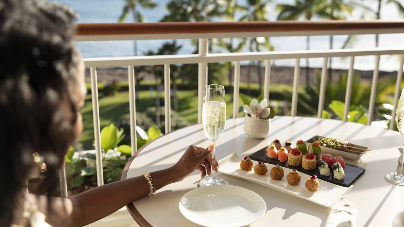 A person holds a champagne flute at a table with three plates of canapes, overlooking a tropical resort and ocean