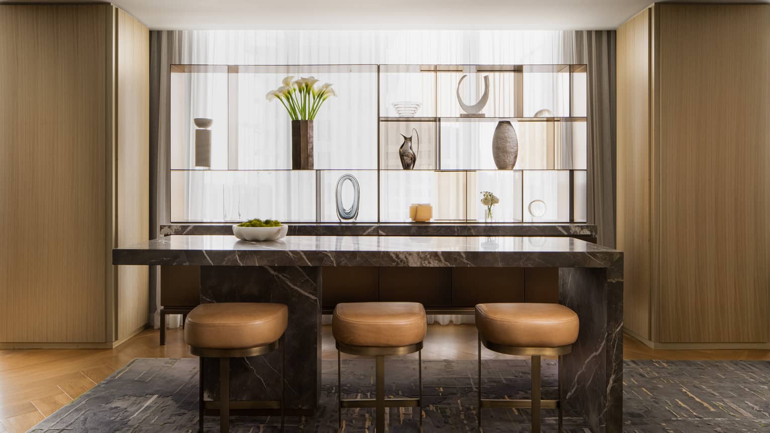 Luxury suite bar area with marble island, three leather stools, and decorative display shelf 