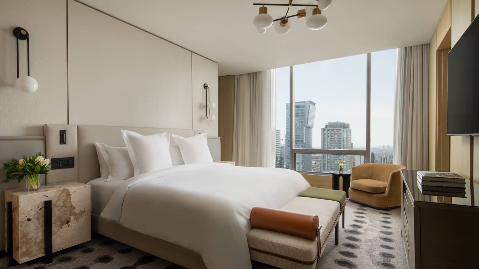 Luxury suite bedroom with a king bed, beige headboard, bench, armchair, TV and floor-to-ceiling windows offering city views