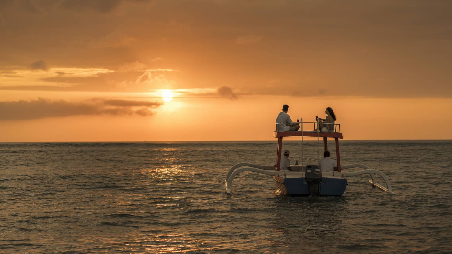 Couple sits on roof of boat sailing on ocean at sunset