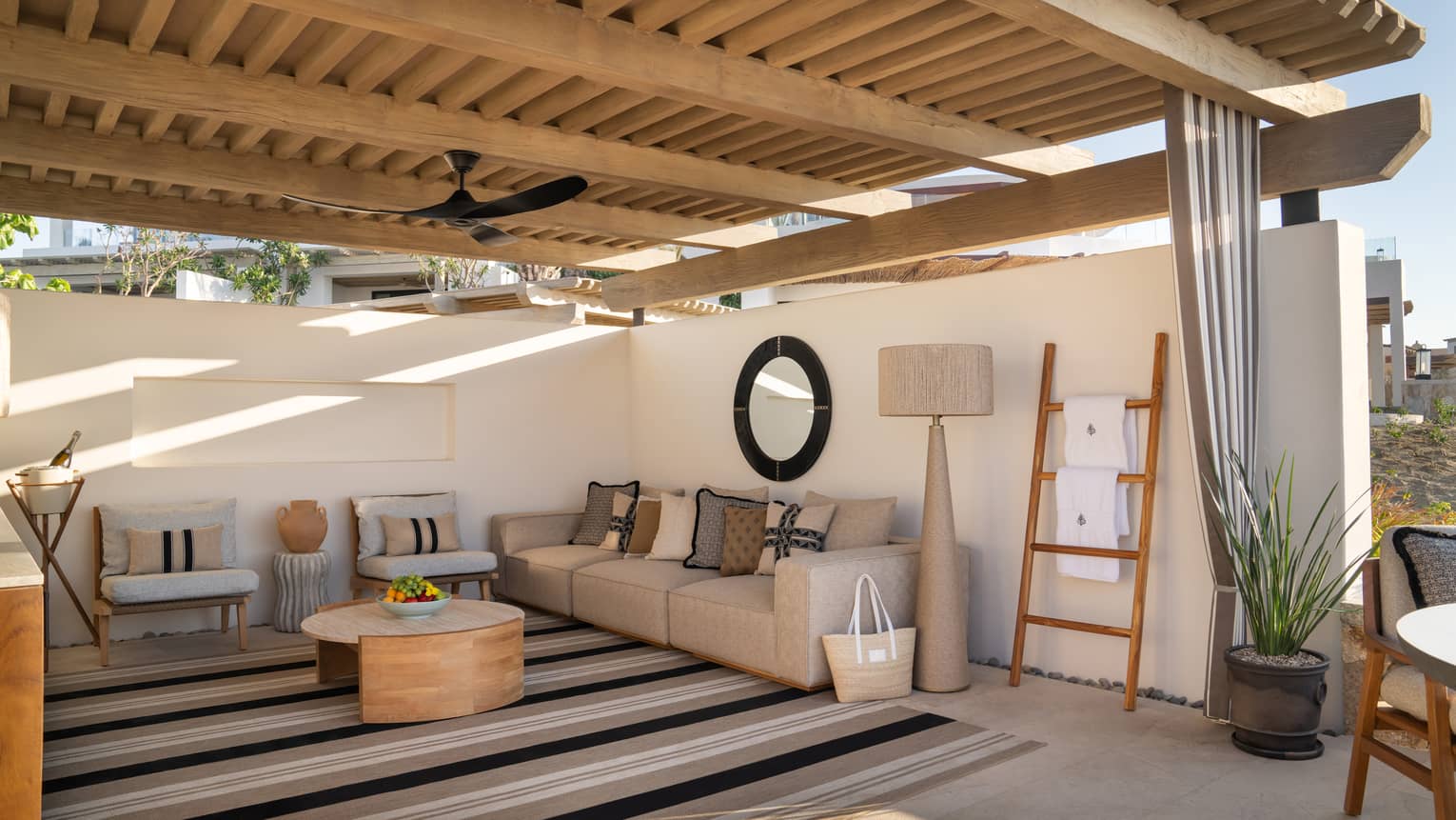 A serene cabana with a raised wooden roof and plaster walls. It's filled with comfortable furniture and a large striped rug.