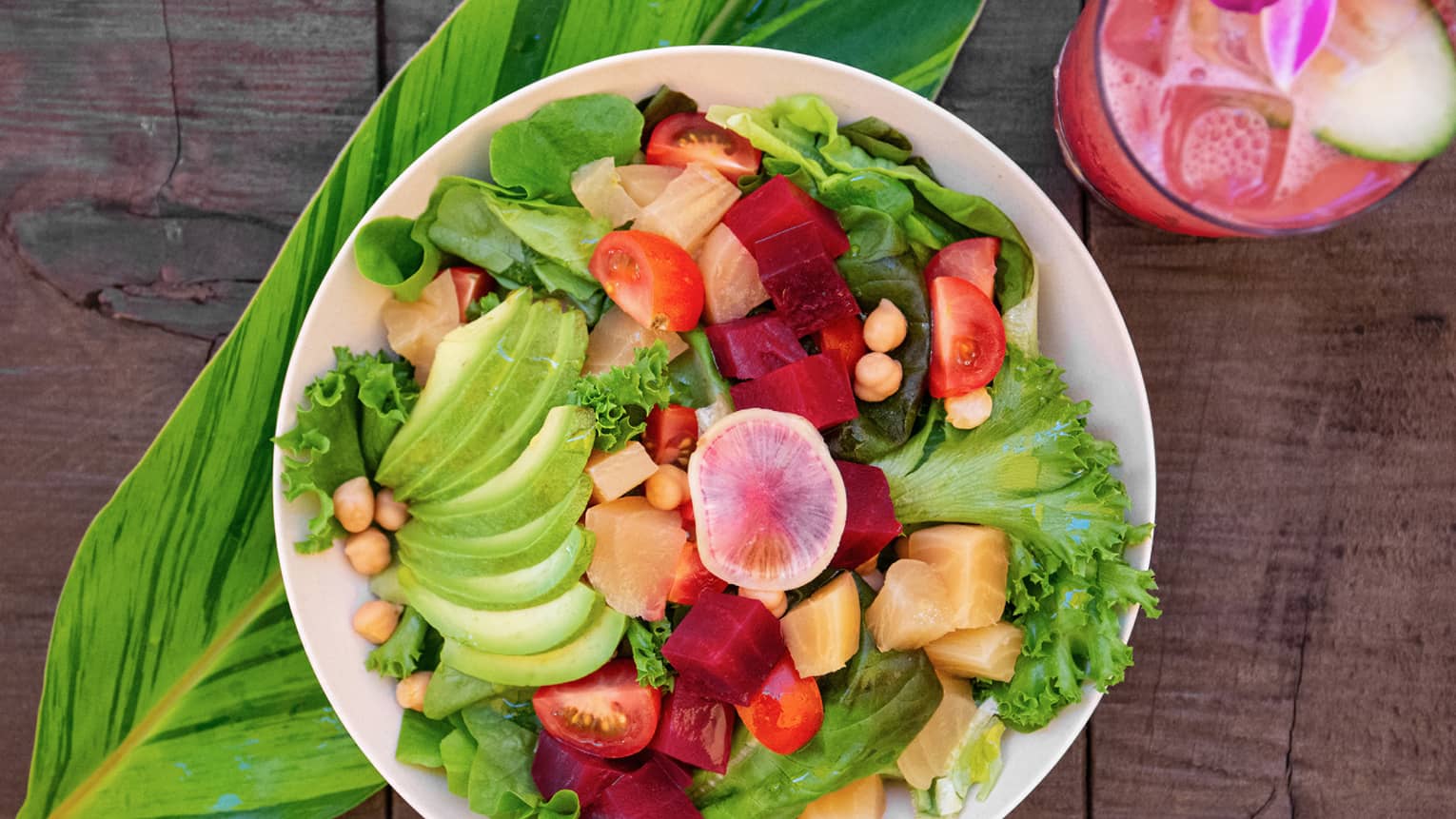 Fresh salad with avocado, tomato and fruit on a wooden table alongside tropical drink
