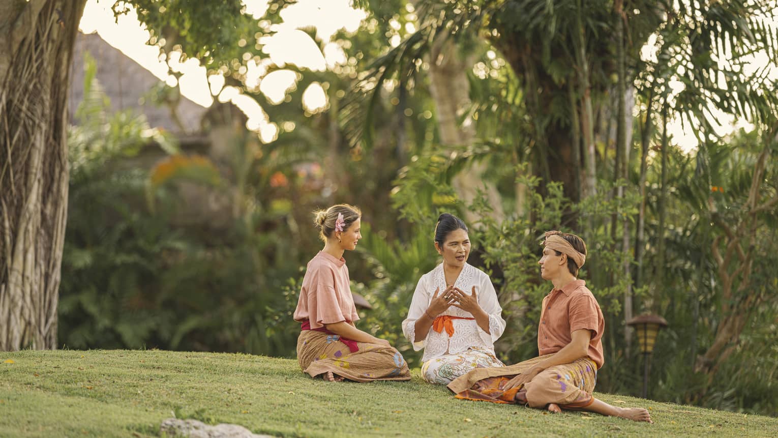 Close-up of three adults in sarongs sitting together on a grassy knoll surrounded by lush greenery and palm trees.