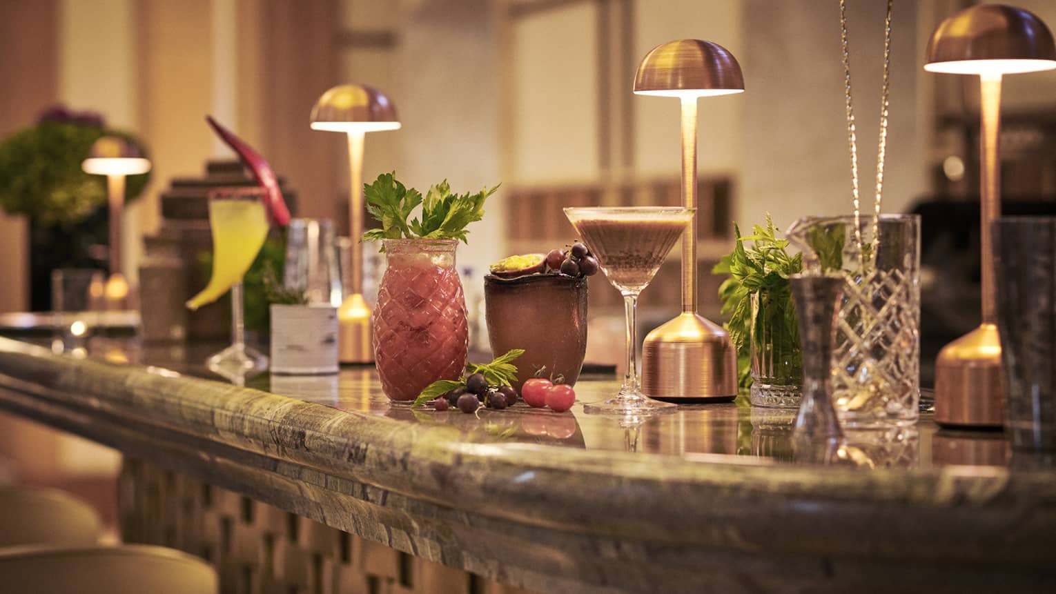 Cocktails in various glassware lined up on bar under copper desk lights