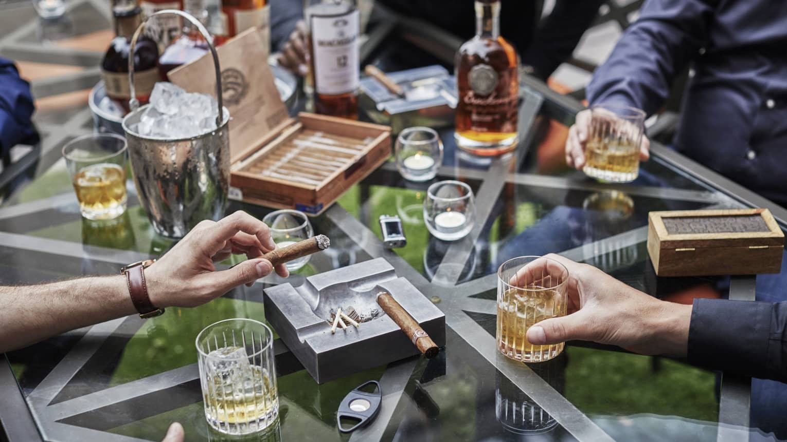 Bachelor party scene with men's hands holding rock glasses with liquor and thick cigars at glass table