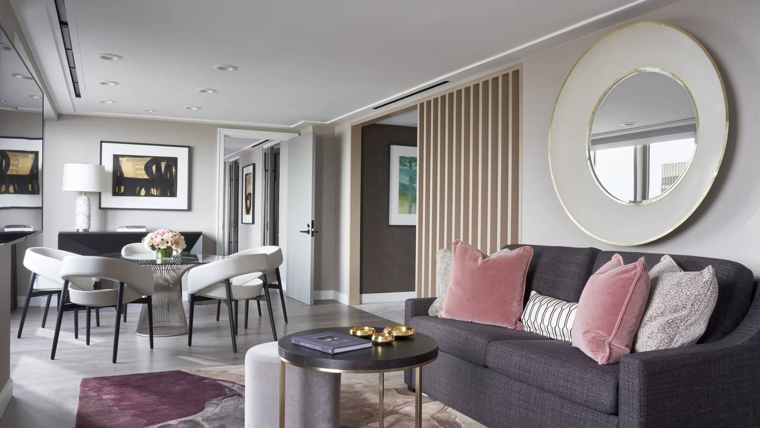 A hotel room with dark grey sofa, pink and grey pillows, coffee table, mirror and small table with chairs.