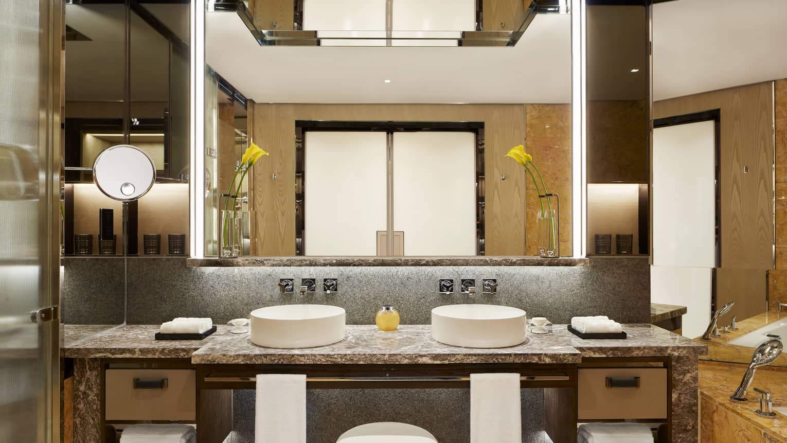 Luxury bathroom with double sinks, marble countertops, large mirror with backlighting and decorative yellow flowers in vases.