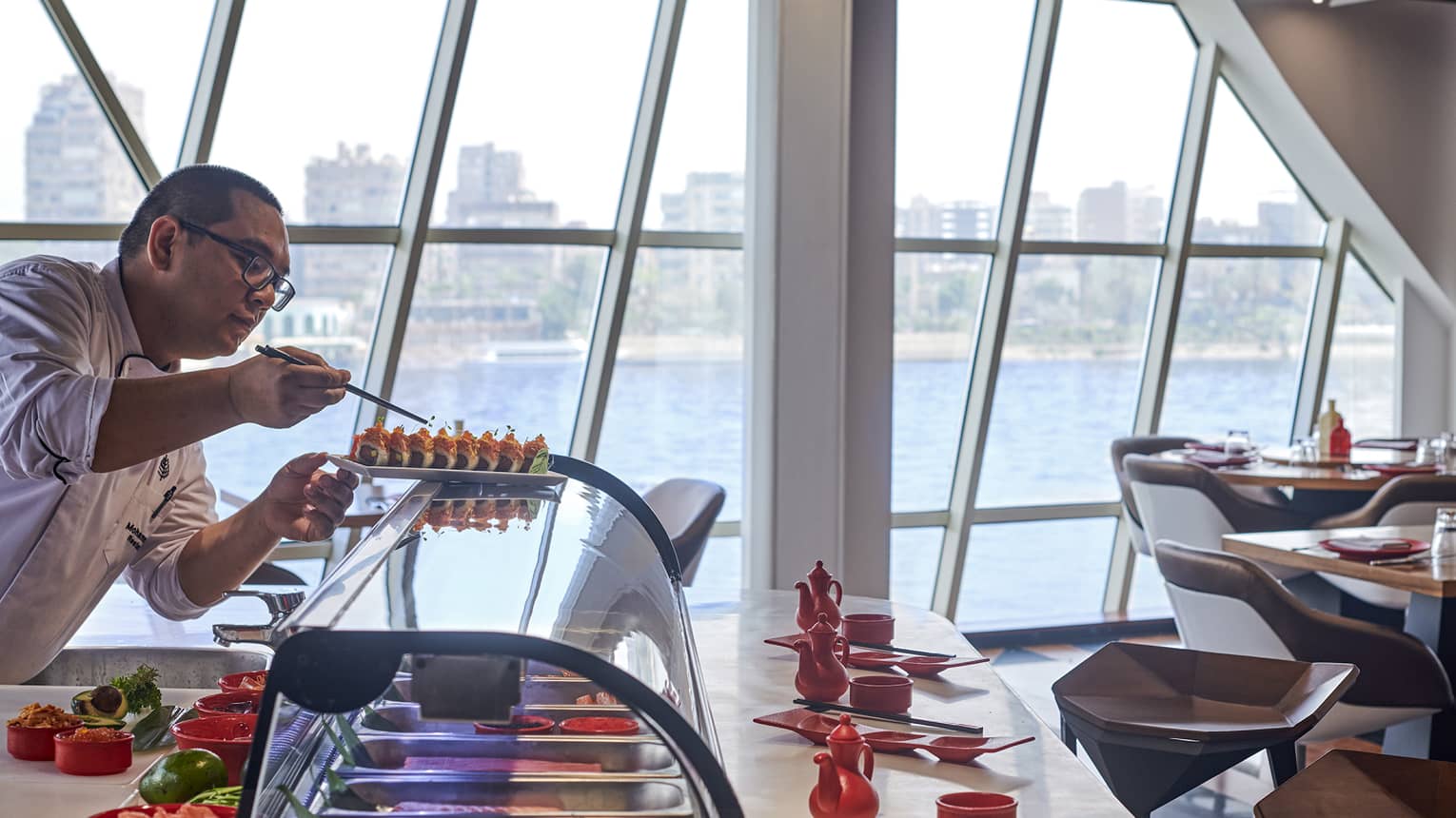 A chef carefully sets tiny greens onto crunchy roll sushi amid bowls, teapots and dipping trays, and panoramic harbour view.