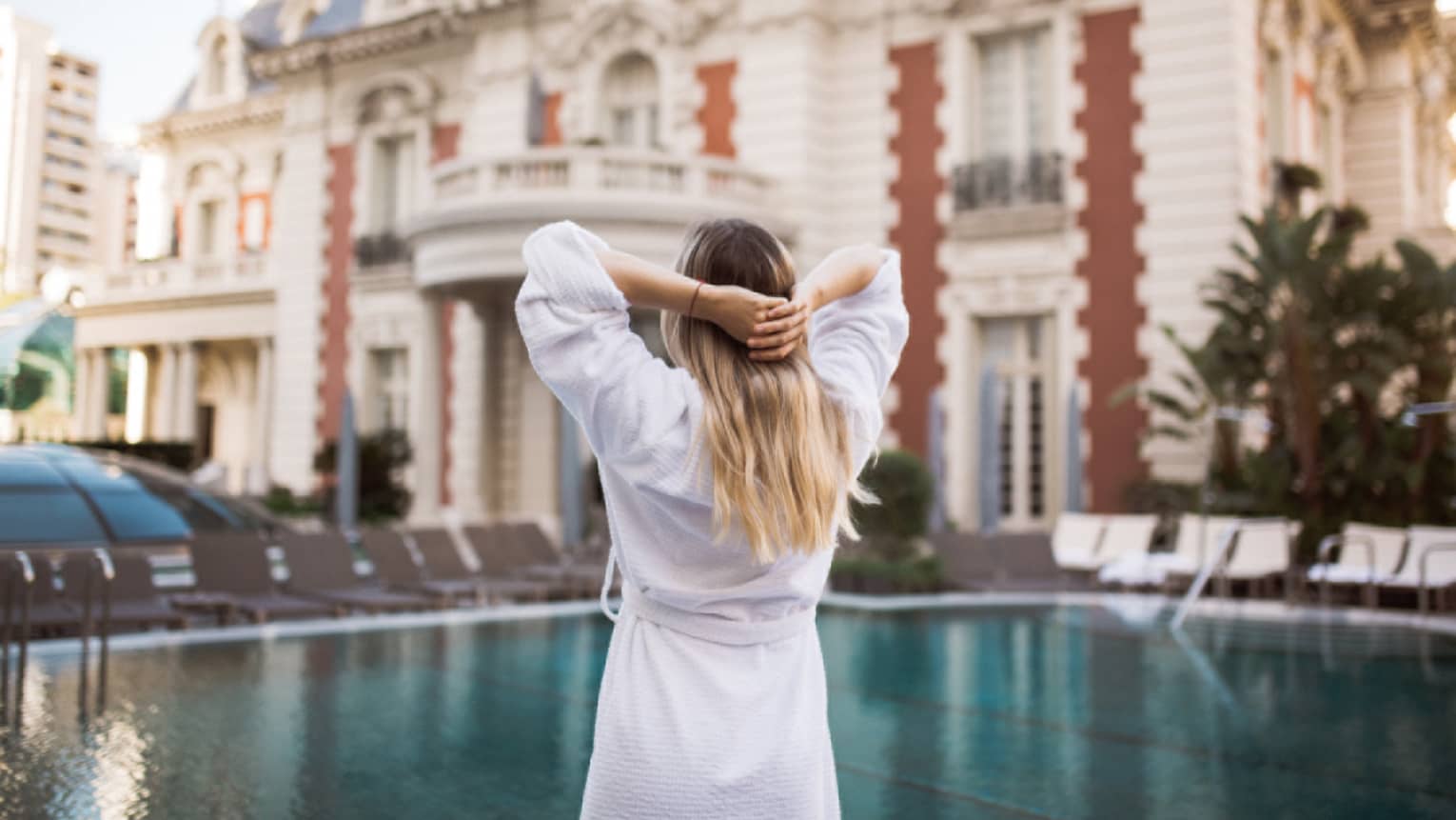 A woman in a white robe stretches by an outdoor pool, the hotel in the background