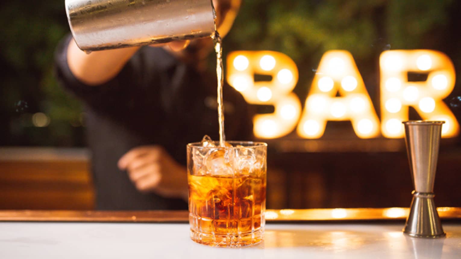 A bartender pours a glass of brown liquor with an illuminated Bar sign in the background