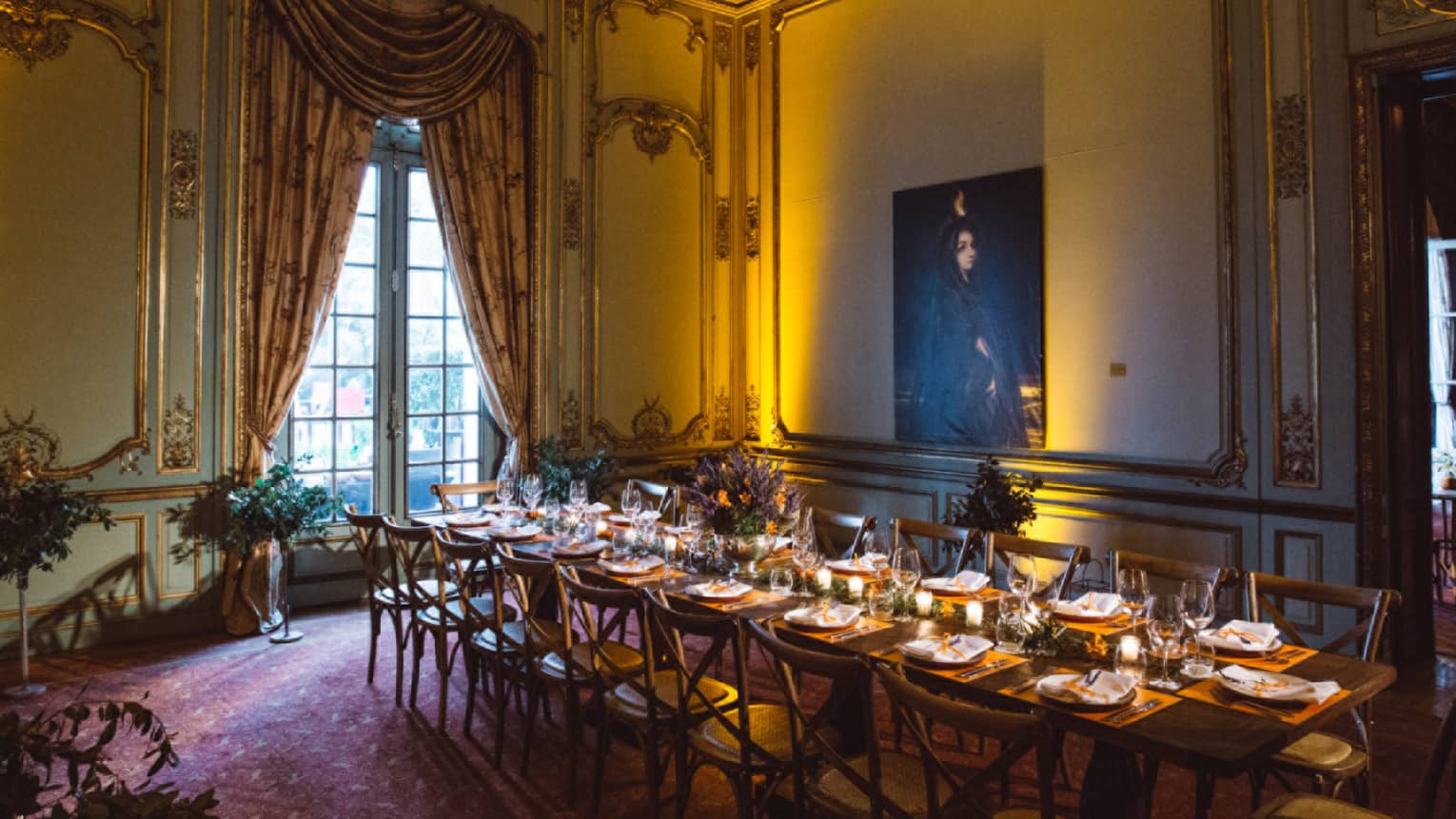 A long rectangular table is set for dinner in a dimly lit dining room with ornate drapes, oriental rugs and a large window