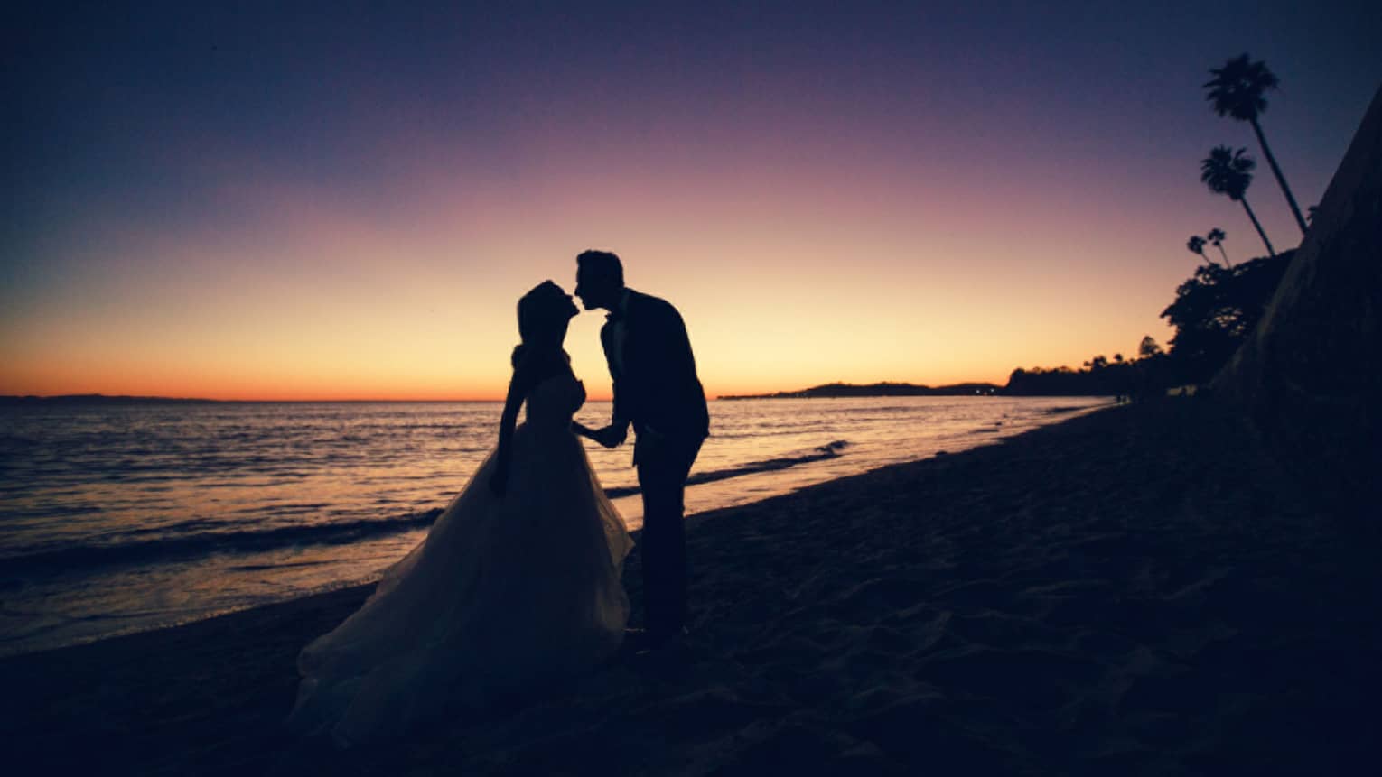Silhouette of bride and groom leaning in for a kiss on beach at sunset
