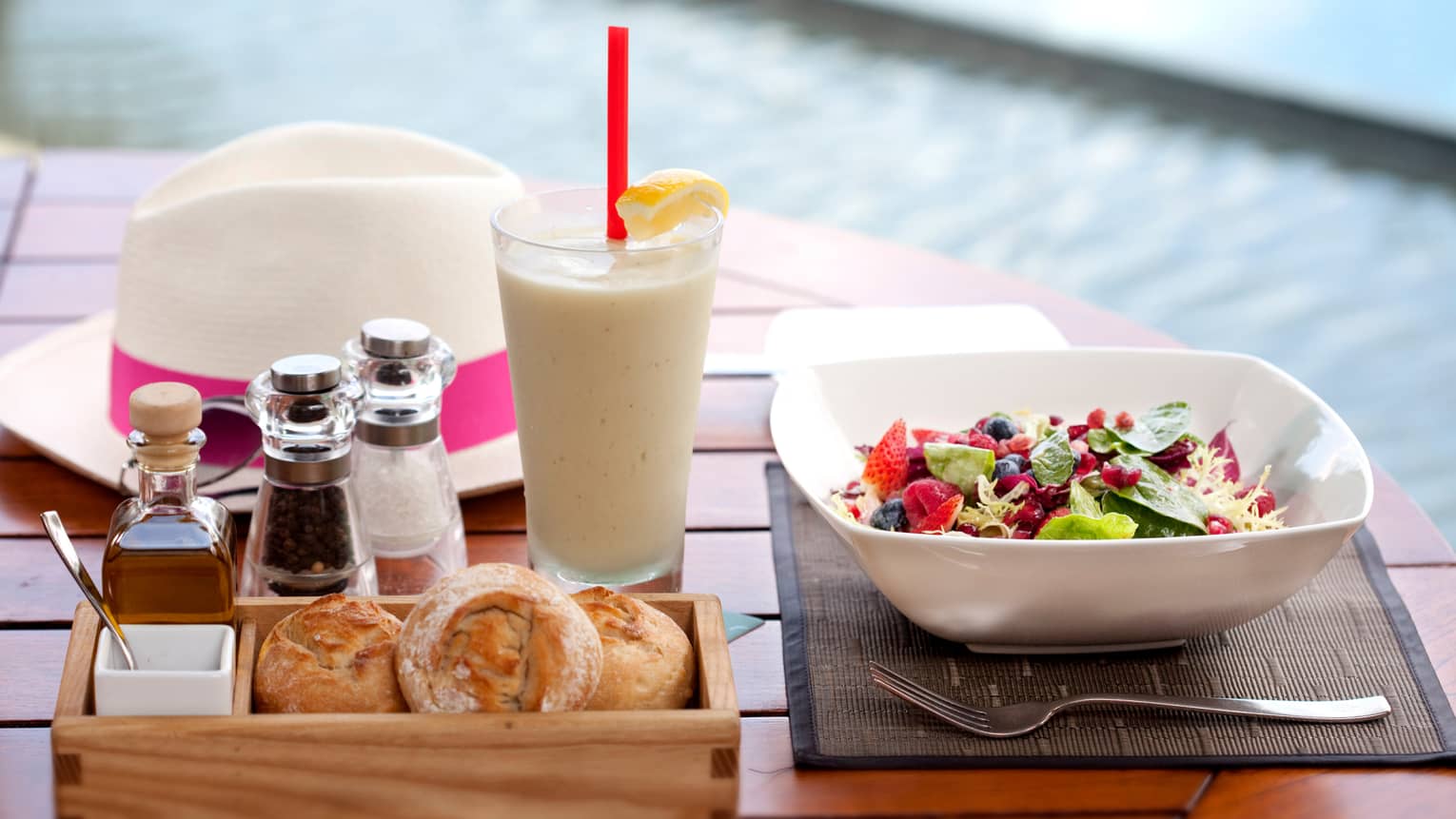 A table by the pool terrace with a meal of mixed salad, bread, condiments, and a cold beverage with lemon. A white hat with a pink band rests in the background.
