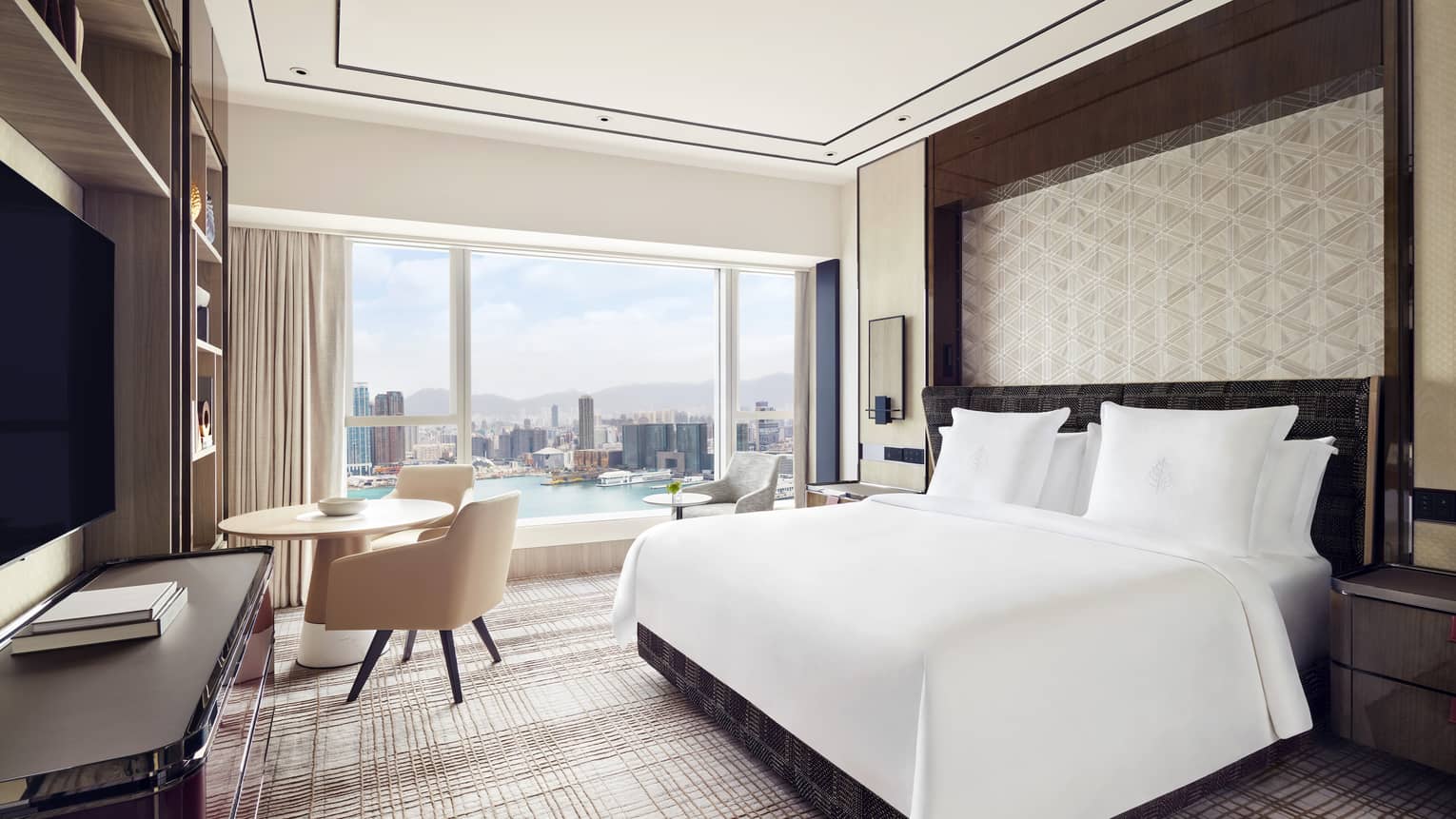 Elegant hotel room with a large bed, patterned accent wall, and a seating area. Floor-to-ceiling windows offer a daytime view of a city skyline and waterfront.