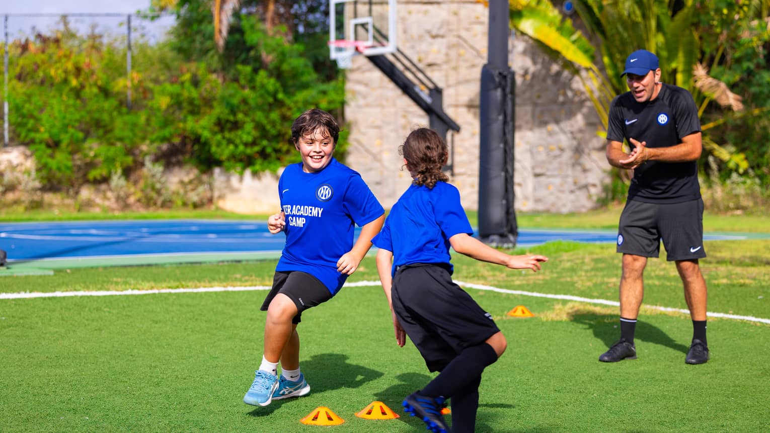 Soccer coach talking to two young guests exercising outside with a basketball court in the background.
