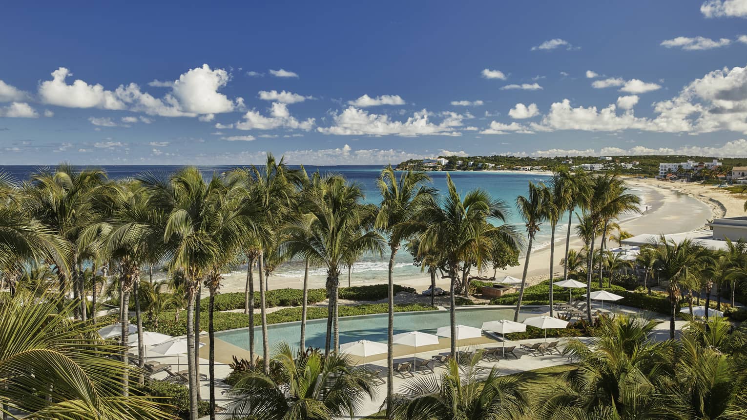 Aerial view of Bamboo Pool surrounded by tall palms, white patio chairs and umbrellas, steps from sand beach and ocean
