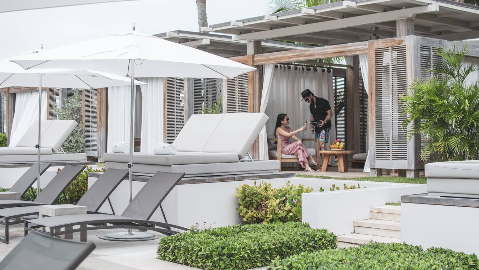 Two talk to each other in a covered pool cabana with a row of white daybeds and a row of grey lounge chairs and white umbrellas in front of them