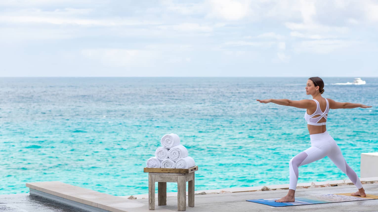 Person practicing yoga in warrior pose by the ocean, next to a stack of rolled towels.