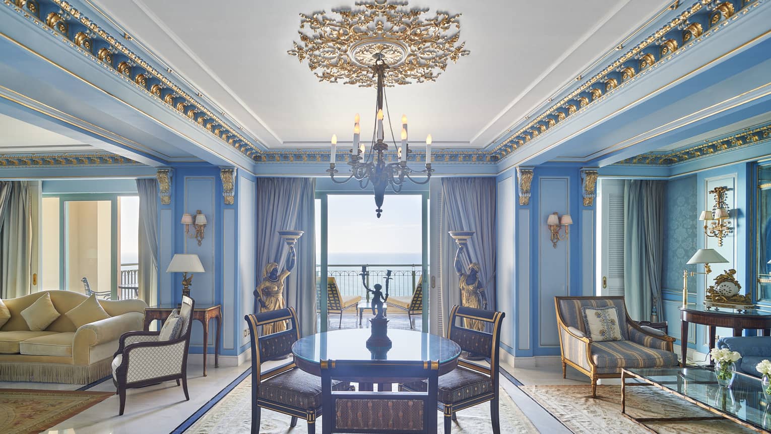 Presidential Suite dining/living area with round dining table, chaise longue, cream couch, blue wall accents, doorway to veranda with sea views