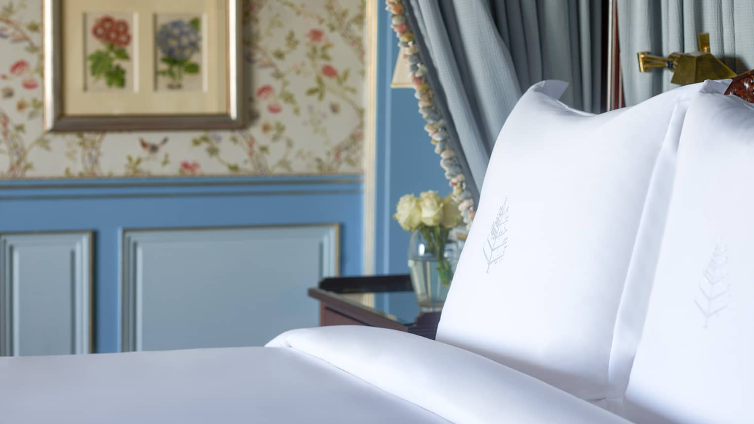 Close-up of bed with white linens, side table with flowers, blue curtains, paisley wallpaper