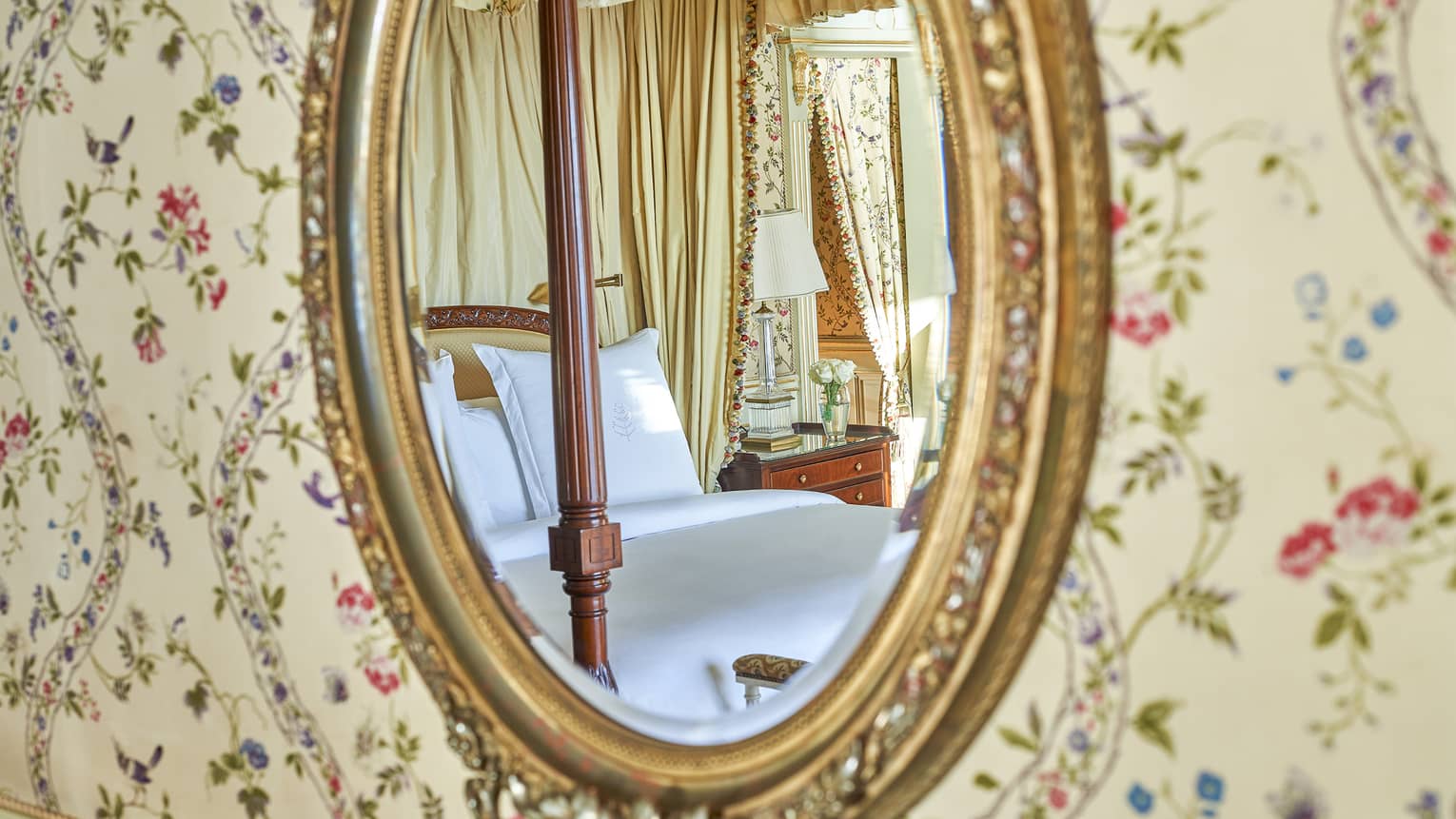 Close-up of oval gold-framed mirror and paisley wallpaper with reflection of bed