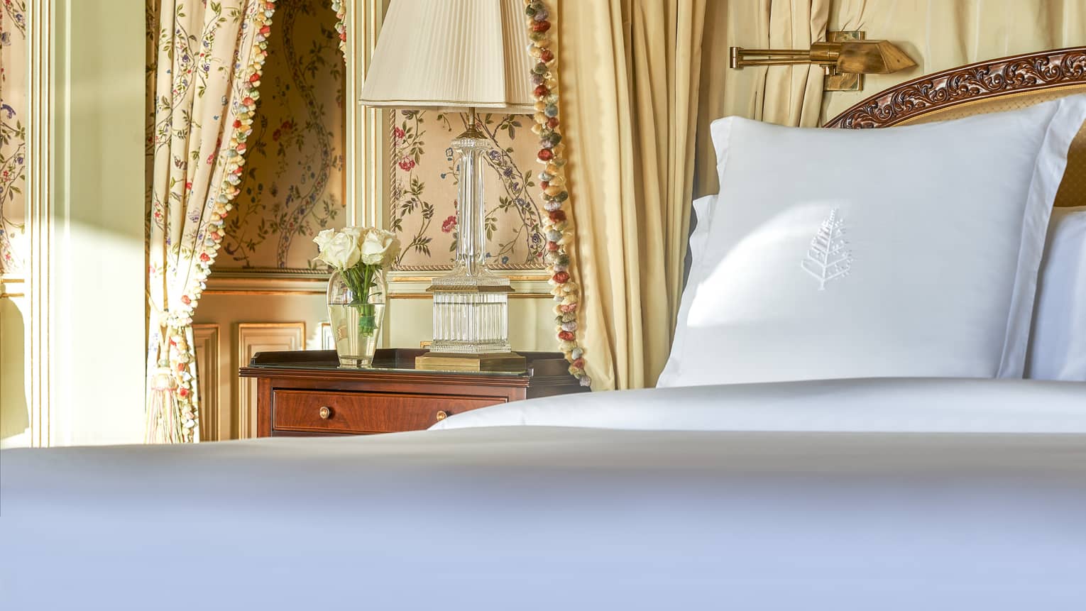 Close-up of bed with white linens, cherry side table, patterned curtains and wallpaper