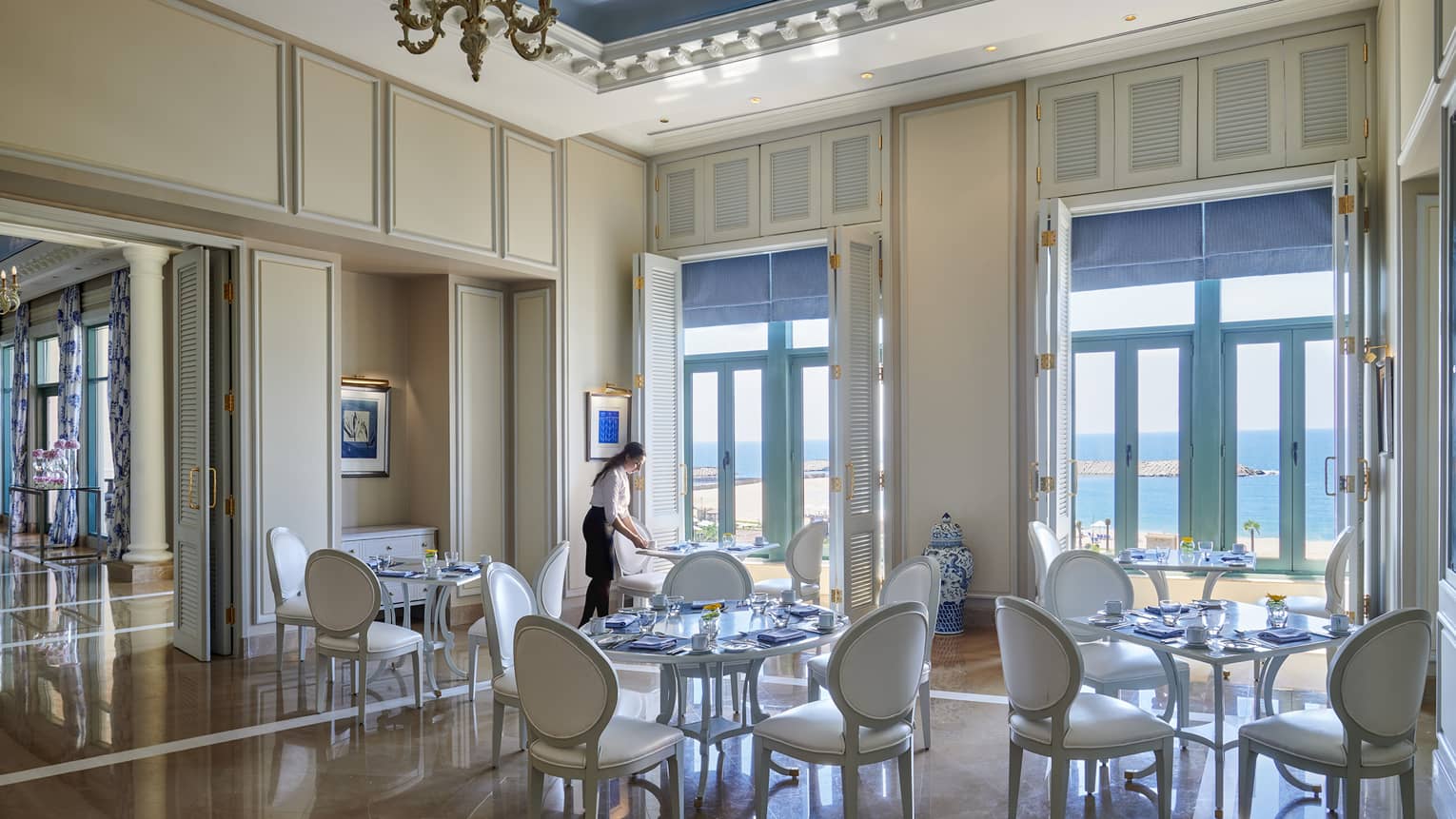 Server sets white table at Kala restaurant, white shutters with blue blinds lead to ocean views 