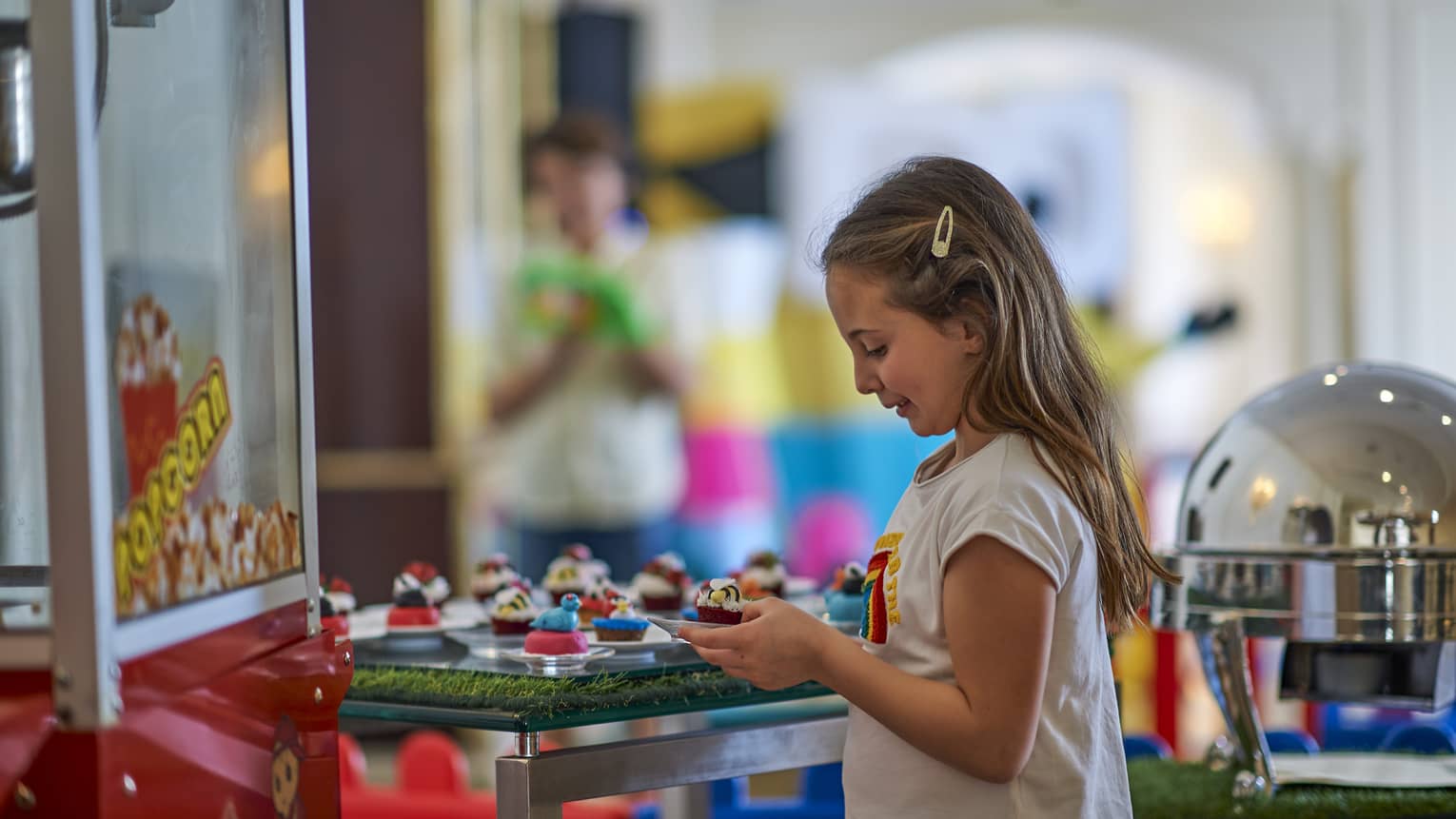 Young girl holds a cupcake at Friday Brunch, a carnival popcorn machine in front of her