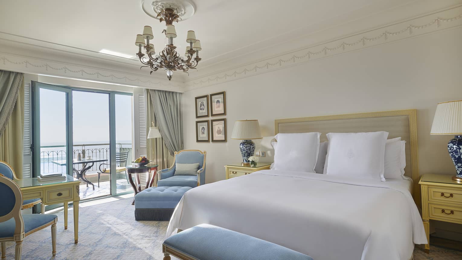 Bedroom with white king bed, chandelier, light blue bench and arm chair, and a balcony overlooking the sea