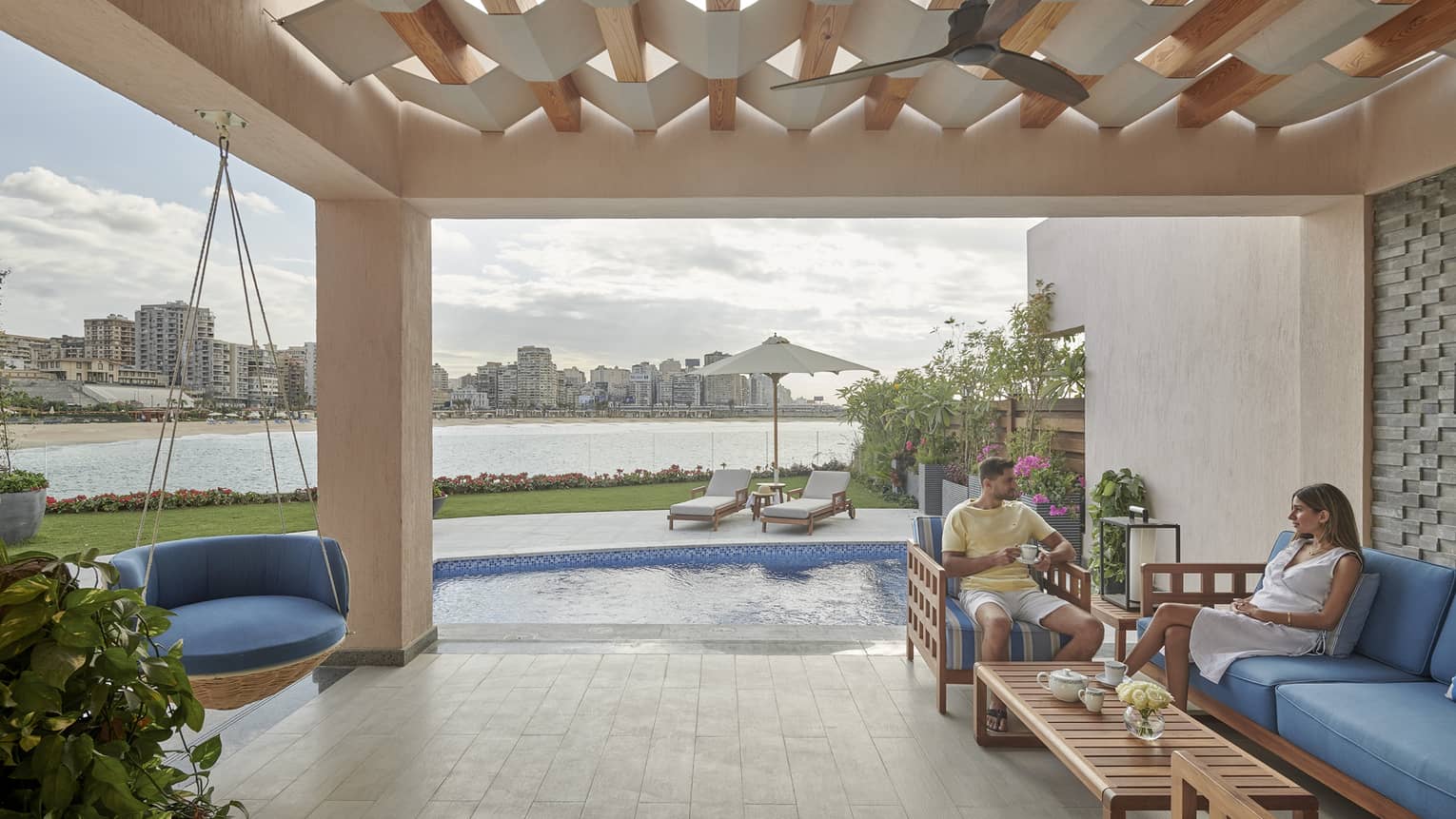 Man and woman sit on blue lounge furniture under pavilion by pool