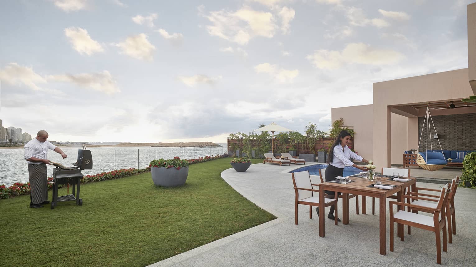 A chef seasons meat on a barbecue near the water’s edge while a server arranges flowers at a table on a patio near a pool.