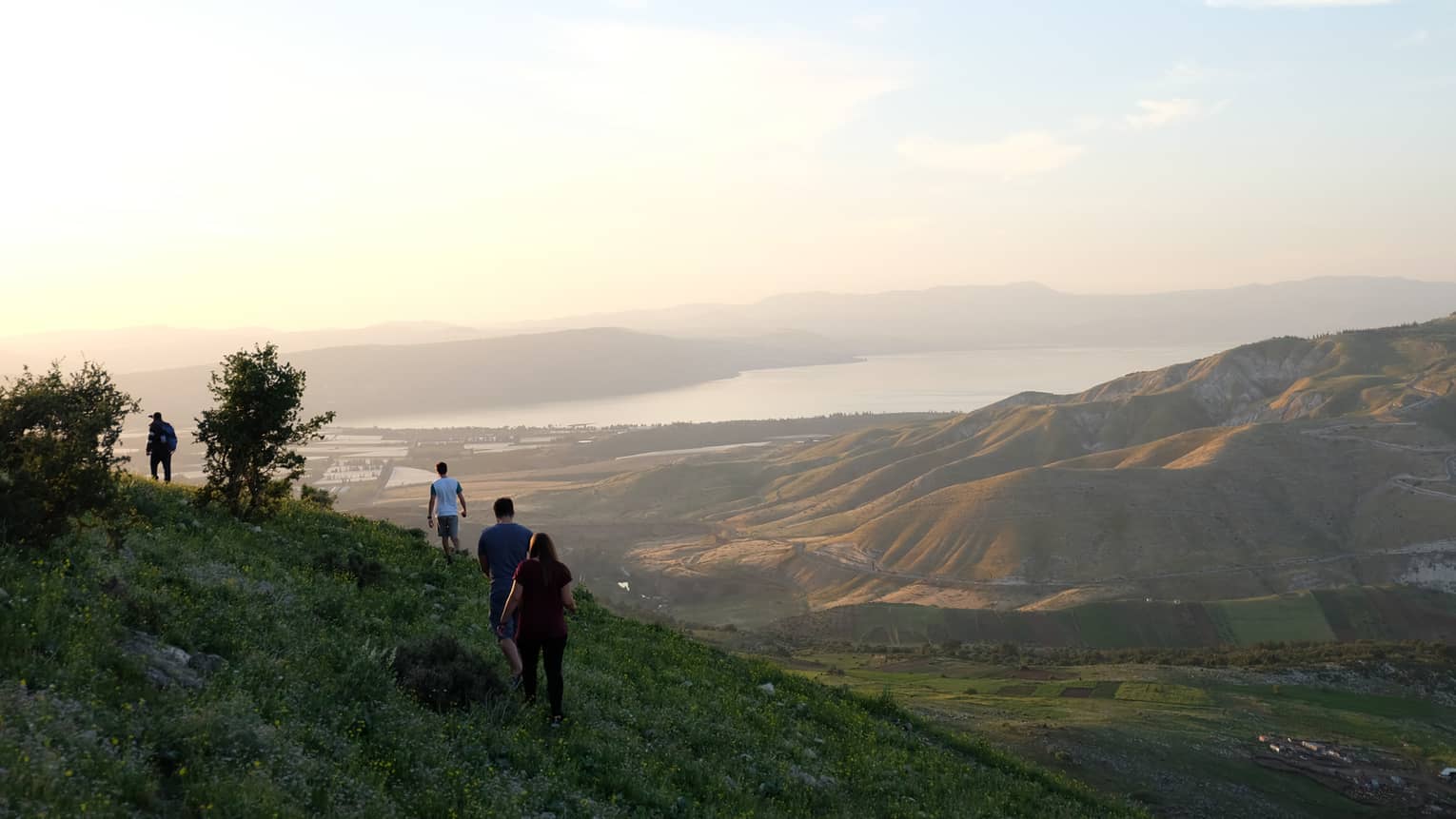 Four people hike in the green, lush, shaded side of a mountain overlooking more mountains and a body of water in Amman 