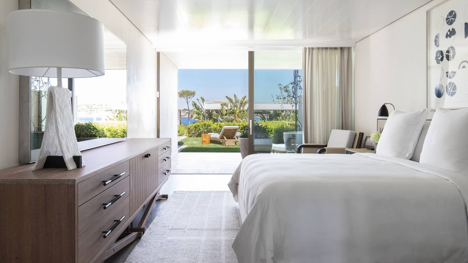Bedroom with plush king bed, modern wooden dresser with lamp, walk-out terrace to garden