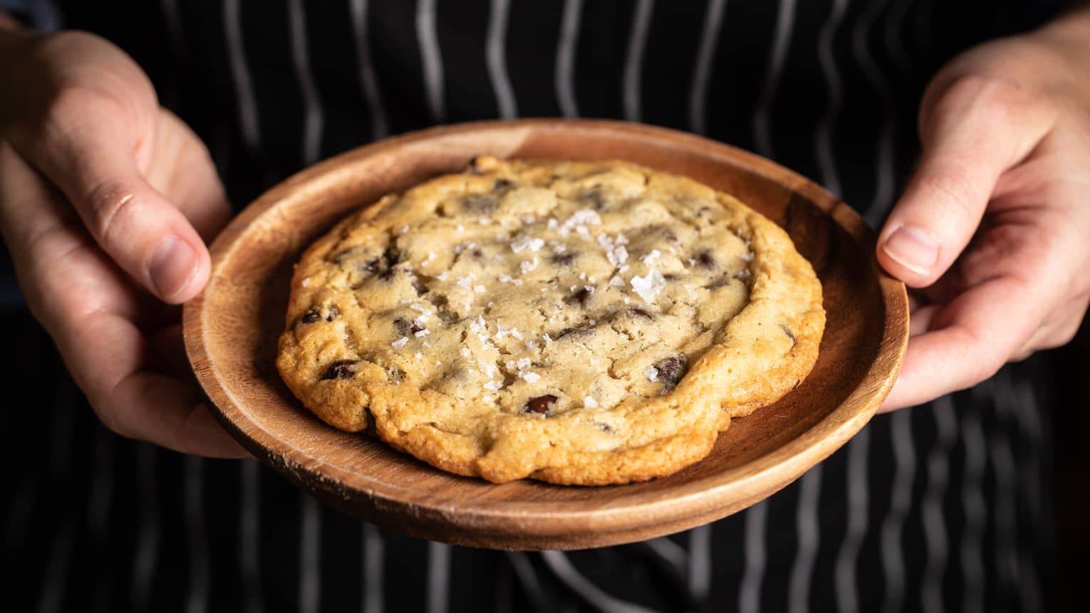 Hands holding a large chocolate chip cookie on a plate.