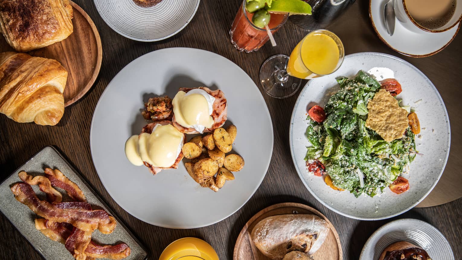 An elaborate brunch, including eggs Benedict, potatoes, bacon, various pastries, salad, a bloody mary and mimosas