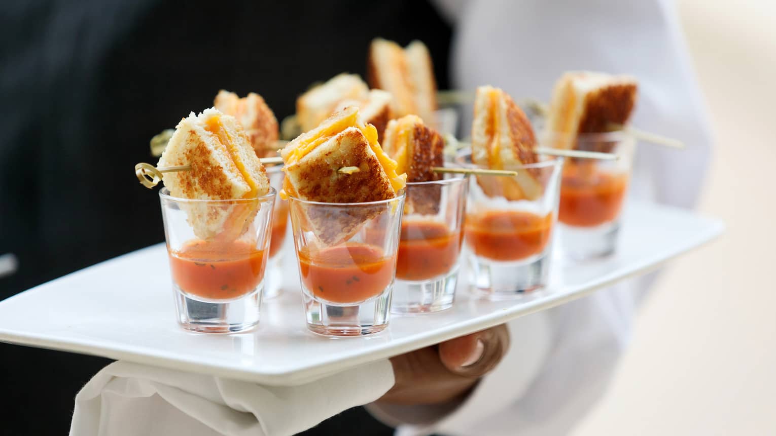Close up of hotel staff holding platter with tomato soup and small grilled cheese sandwiches in shot glasses