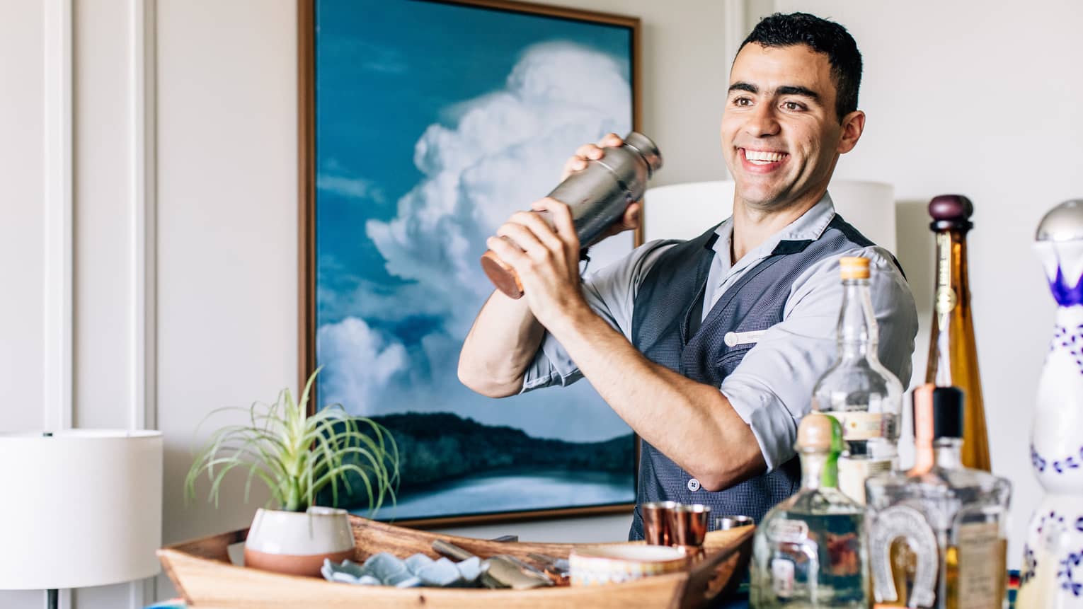 A bartender shaking a cocktail with liquor bottles on a cart in front of him.