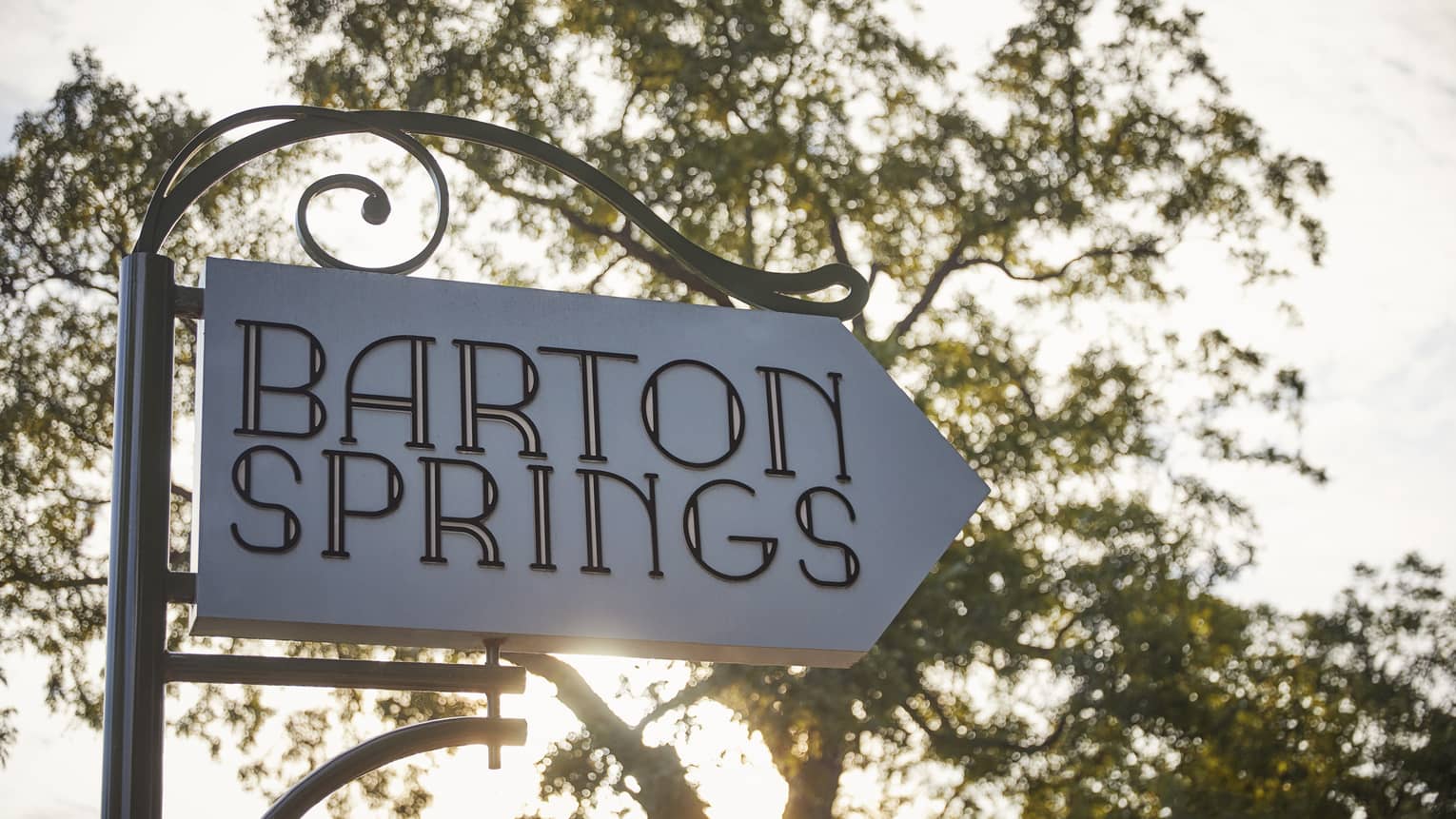 A white sign with a large tree and the sun behind it that reads "Barton Springs"