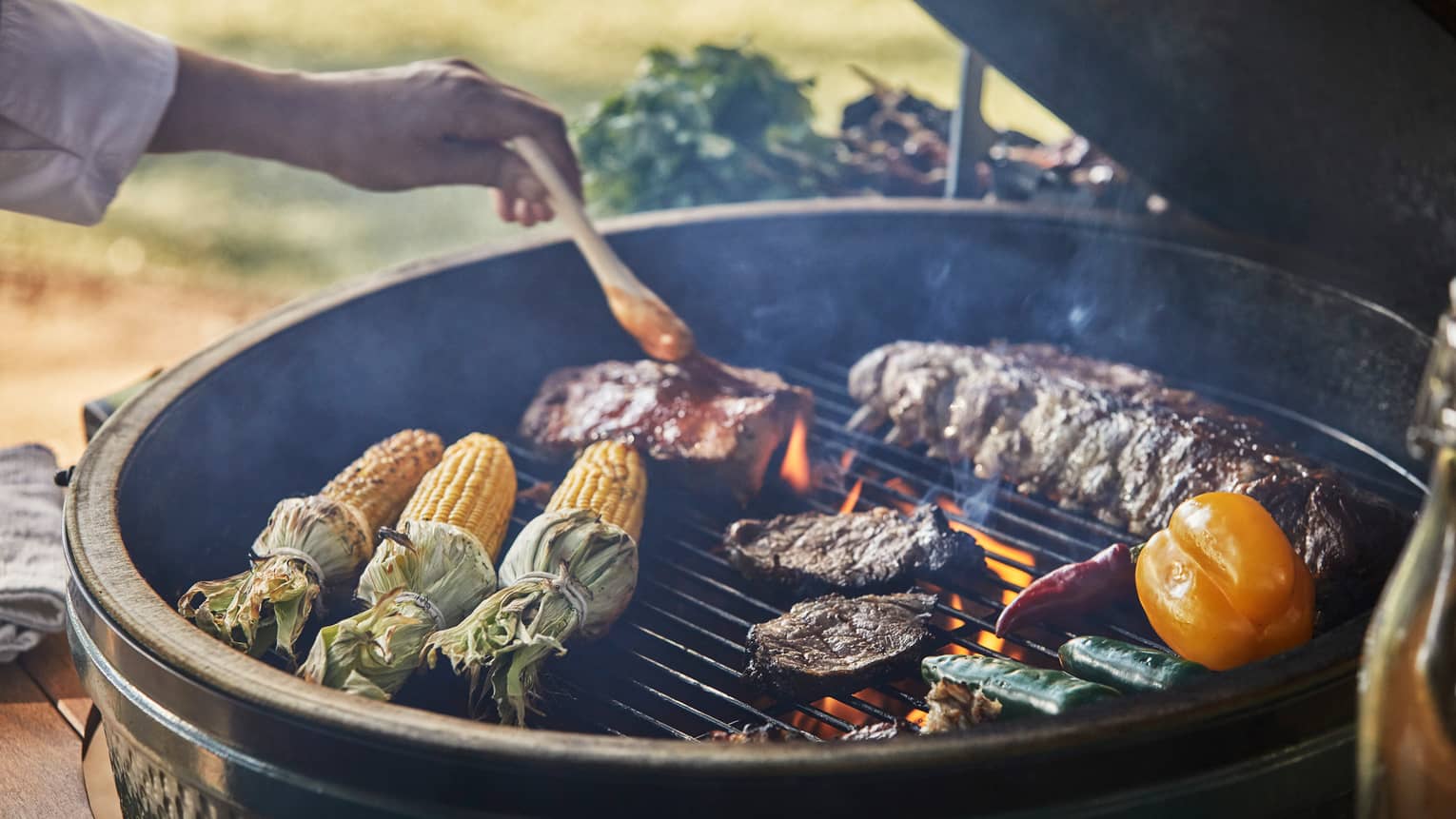 Close-up of chef brushing sauce on ribs on outdoor barbecue with corn, steaks, vegetables