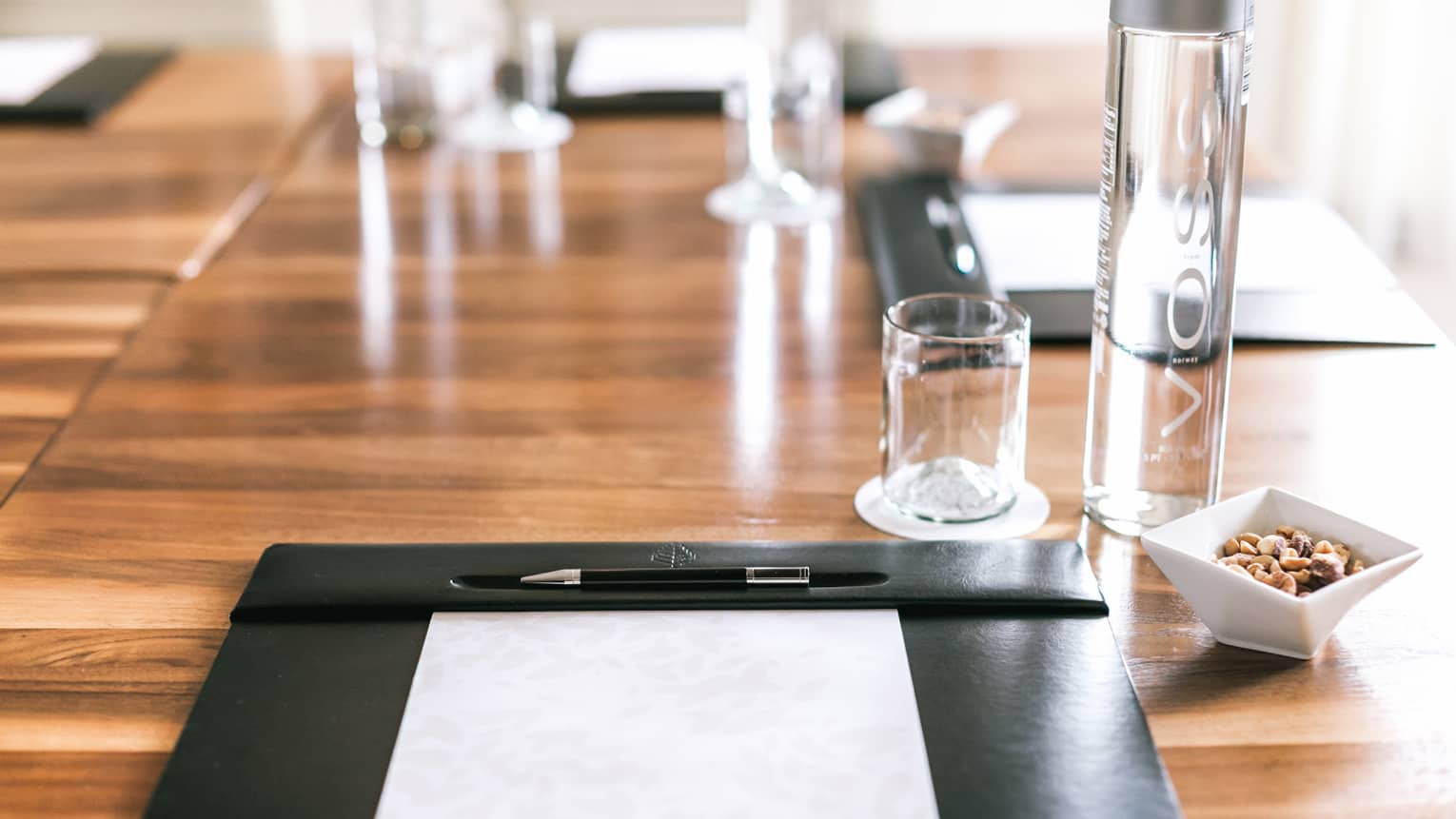Close-up of meeting table with leather agenda and pen, glass VOSS water bottle, bowl with nuts