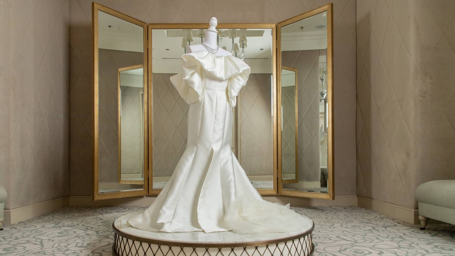 A wedding dress on a pedestal in front of a 3-part mirror.