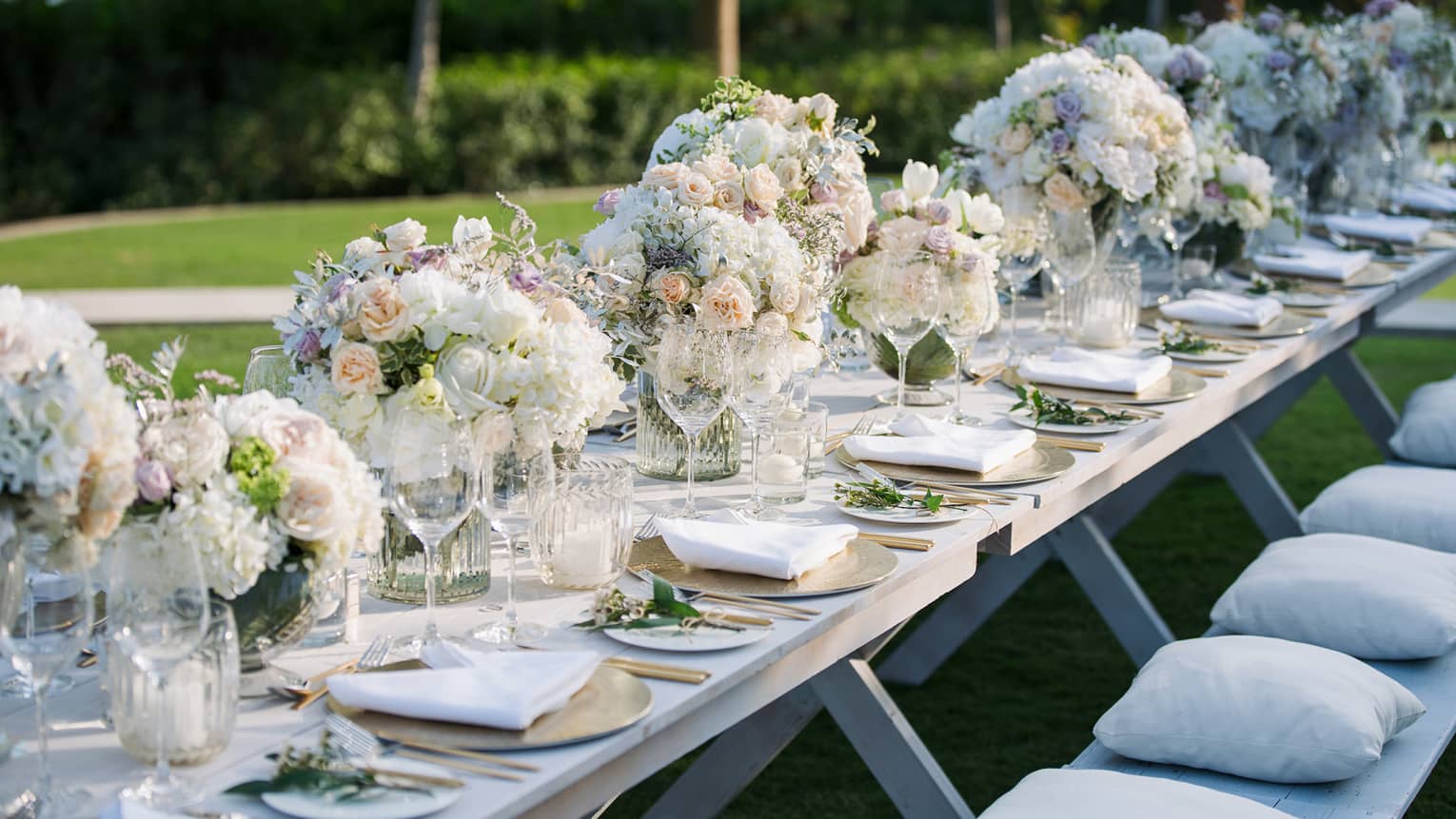 A banquet style table is set with gold chargers, white plates, tableware and large white bouqets