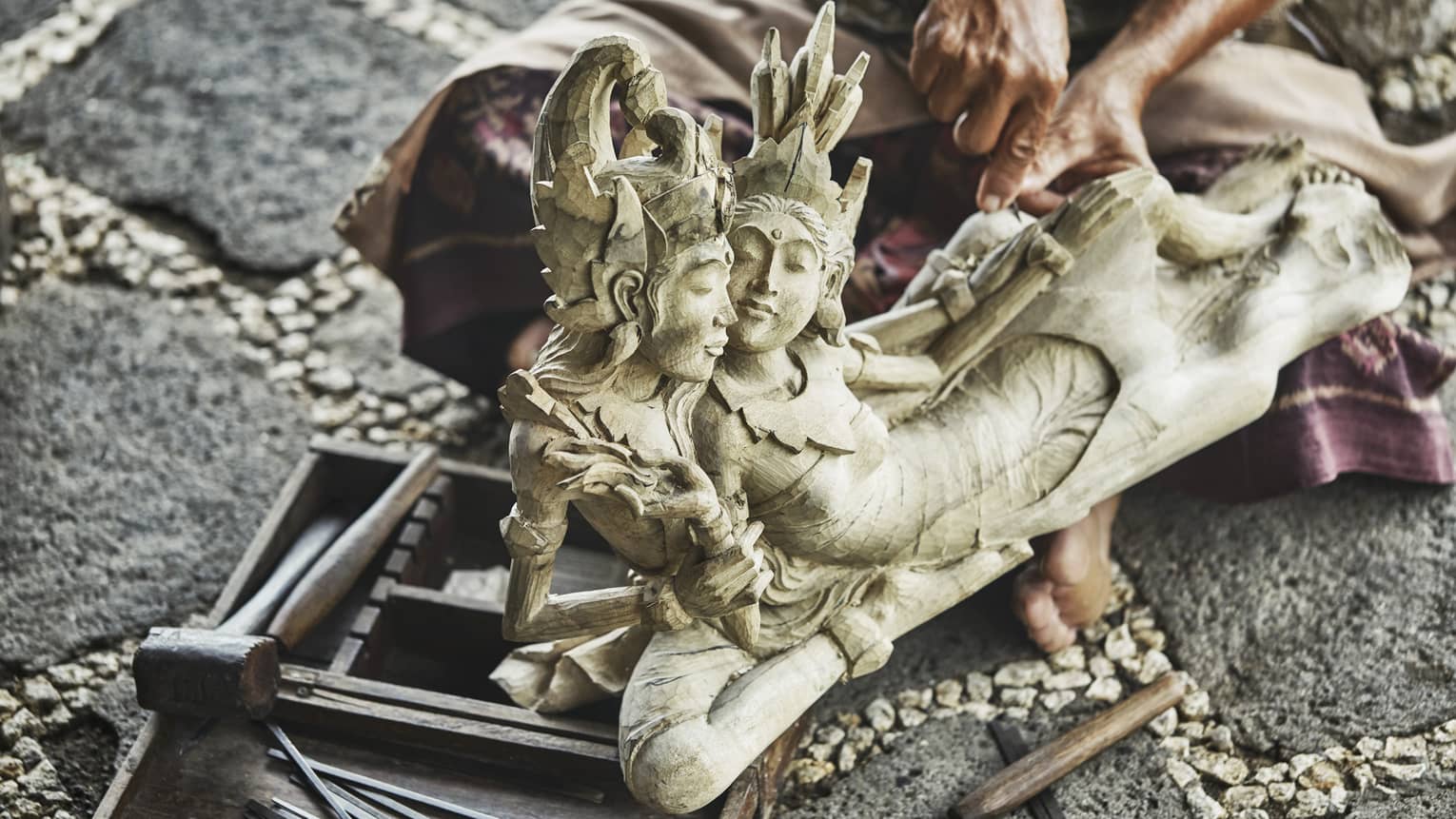 Close-up of intricate carved wood statue, man's hands with carving knife visible behind