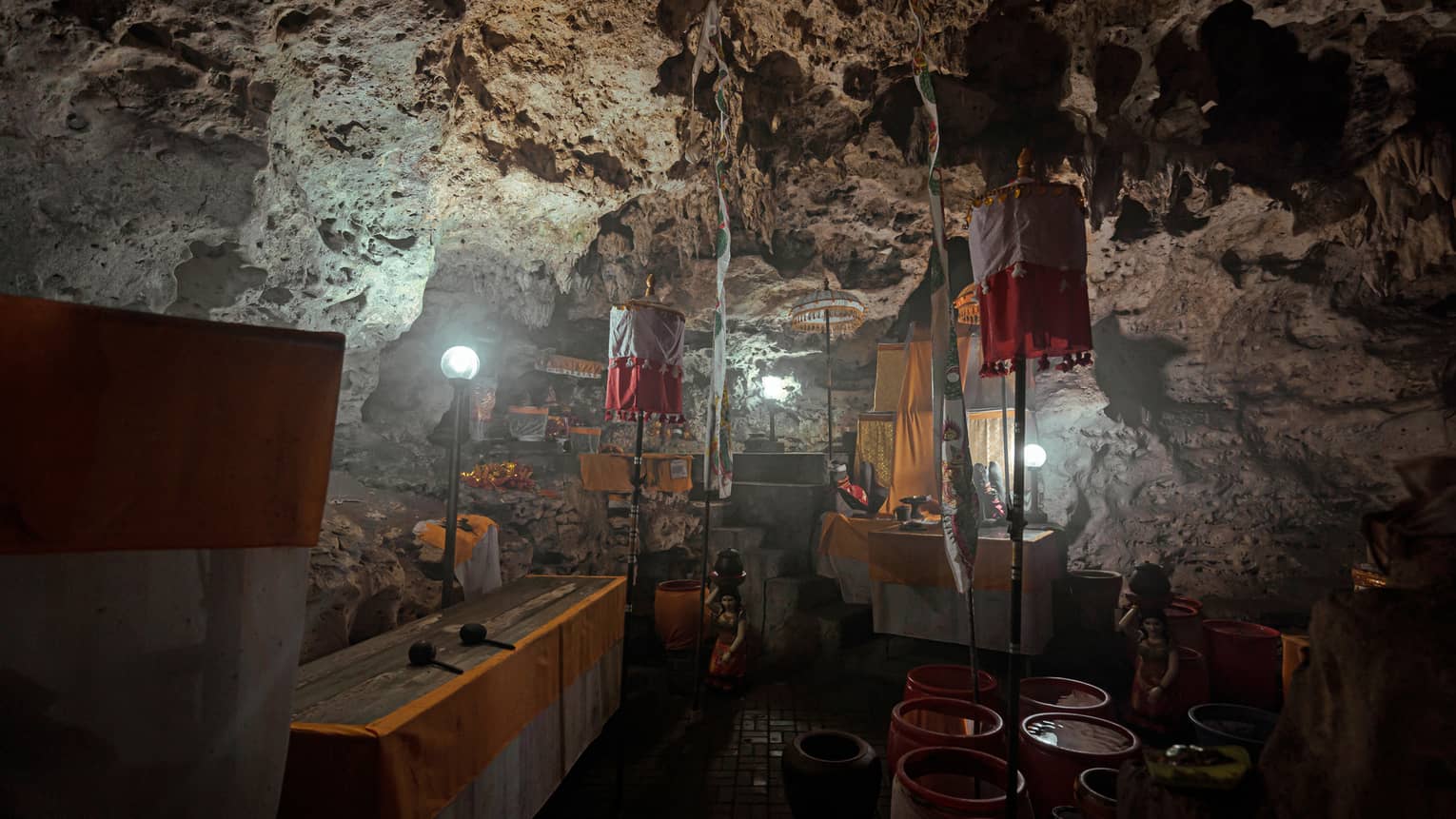 A temple inside a dim, rocky cave lit by lamps and adorned with pots, statues, wooden tables and hanging yellow fabrics.