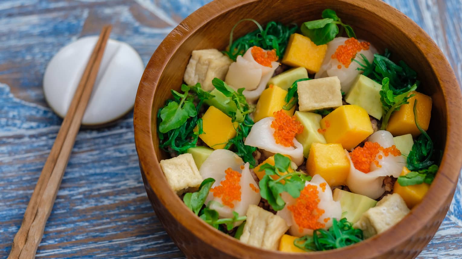 A wooden bowl full of colourful veggies and tofu, with wooden chopsticks resting on a porcelain dish beside it
