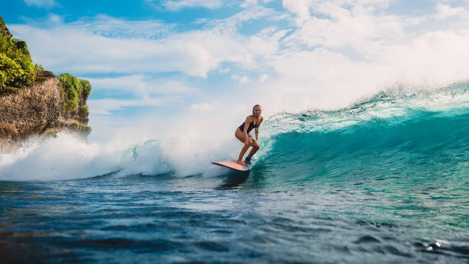 Under blue skies, a blond surfer with knees bent and eyes straight ahead rides a frothy wave, passing by a large mossy cliff.