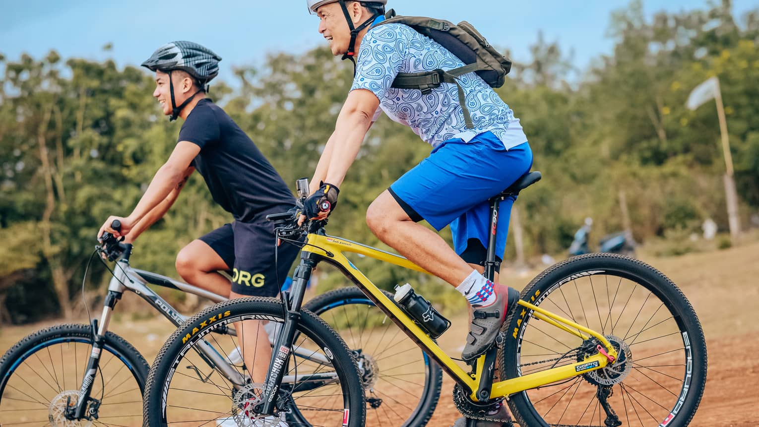 Side view of two smiling mountain bikers in helmets as they coast down a gentle slope on a dirt path alongside trees.