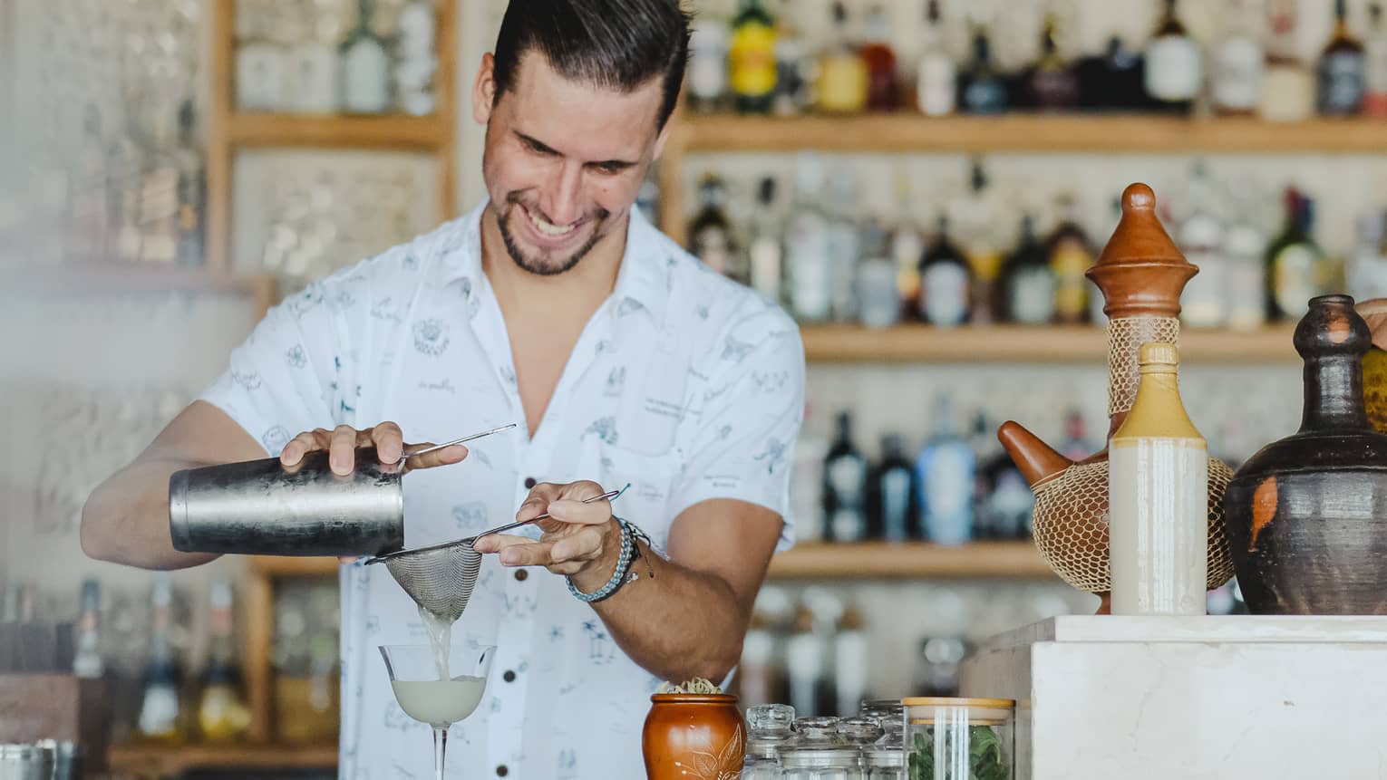 Close-up of a smiling bartender pouring a cloudy liquid from a metal cocktail shaker through a strainer into a stemmed glass.