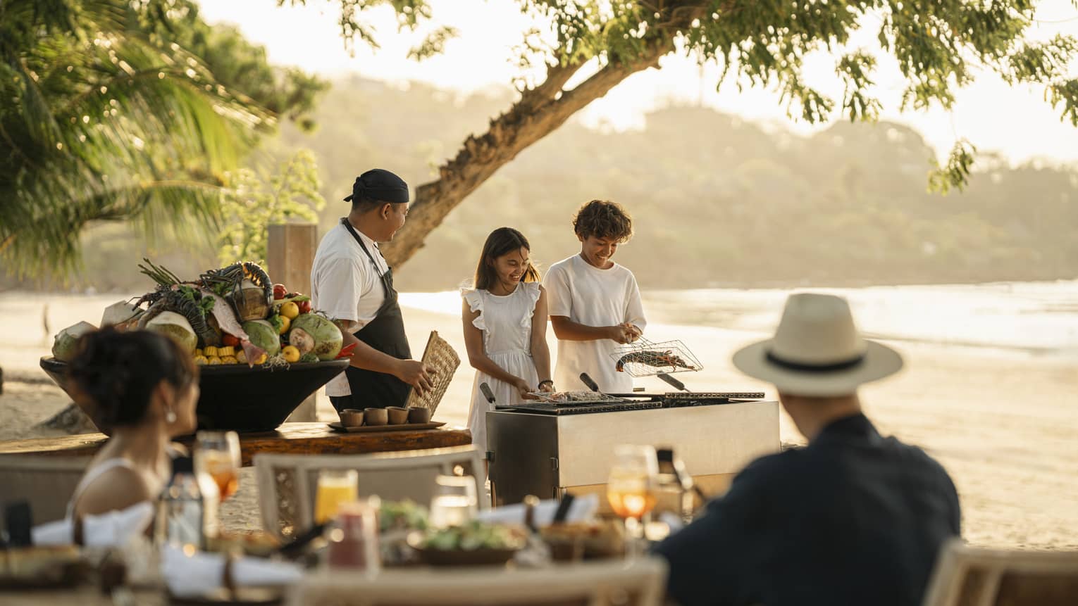 A chef smiles at two children who barbecue shellfish in a grilling basket on the beach as their parents watch.