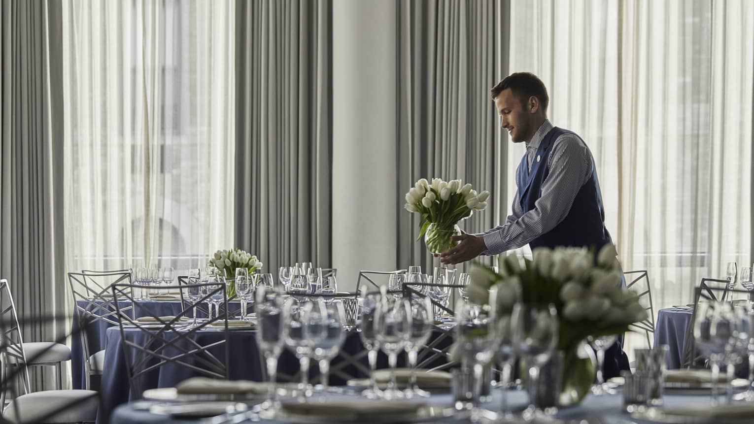A four seasons male staff placing a white floral arrangement onto a circular table, draped in a dark gray table-cloth